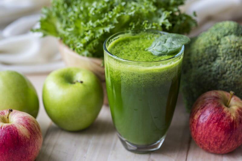 Glass of green vegetable juice surrounded by apples, broccoli and leafy greens on a wooden table, fresh and healthy mood