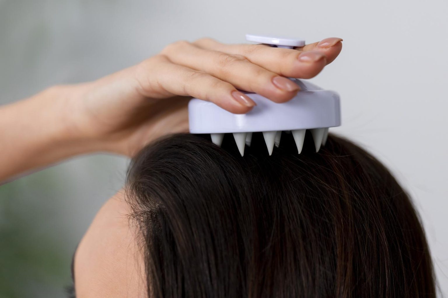 Close-up of a hand holding a light purple scalp massage brush on dark hair, soft neutral background, calm mood